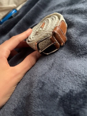 Beige flätad bälte med läderdetaljer - Säljer ett beige flätat bälte med snygga bruna läderdetaljer och silverfärgat spänne. Bältet är ca 110 cm långt och passar perfekt till jeans eller chinos för en clean och stilren look.