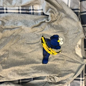 Grå sweatshirt med blå figur och banan klaws Uniqlo - Säljer en grå sweatshirt från Uniqlo med en stor blå tecknad figur och en gul banan på bröstet i samarbete med Klaws. Tröjan har rund hals och långa ärmar, perfekt för dig som gillar streetwear och coola prints.