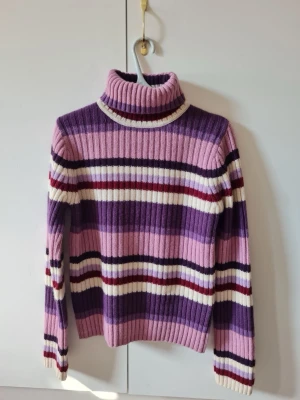 Varm ulltröja från Åhléns strl L - Säljer en ribbstickad polotröja retro från Åhléns i storlek L, gjord av 70% lammull och 30% akryl. Tröjan har breda ränder i lila, rosa, vinröd och vit, samt långärmad design och hög krage.