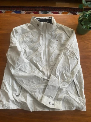 Vit vindjacka från Tommy Hilfiger - Säljer en stilren vit vindjacka från Tommy Hilfiger med diskret logga på bröstet. Jackan har hög krage, dragkedja framtill och två sidofickor med dragkedja. Insidan är meshfodrad för extra komfort och jackan har en sportig look. Storleken är XL men känns mer som en L