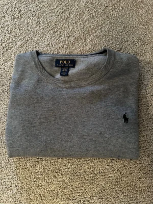 Grå stickad tröja från Polo Ralph Lauren - Klassisk grå stickad tröja från Polo Ralph Lauren med rund halsringning och diskret broderad logga på bröstet. Tröjan har ribbade muddar vid ärmslut och nederkant, perfekt för lager på lager under höst och vinter. Är storlek xl barn men sitter som en vuxen S