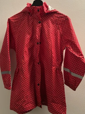 Röd prickig regnjacka med huva barnstorlek - Snygg röd regnjacka med vita prickar och reflexdetaljer på ärmarna. Jackan har huva, både knappar och dragkedja framtill. 