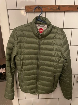 Olivgrön pufferjacka från Colmar - Säljer en olivgrön pufferjacka från Colmar med quiltad design och hög krage. Jackan har dragkedja framtill och två sidofickor med dragkedja. Insidan är ljusgrå och materialet är syntet. Perfekt för kyliga dagar!