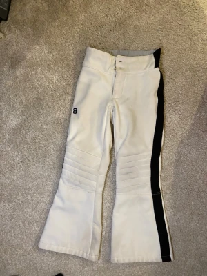 Vita täckbyxor från 8848 Altitude - Säljer ett par vita täckbyxor från 8848 Altitude med svart sidopanel och broderad logga på benet. Byxorna har hög midja, dubbla knappar och mjukt fleecefoder på insidan. Snygga sömdetaljer över knäna och bootcut passform. Perfekta att ha när man åker skidor⛷️😍