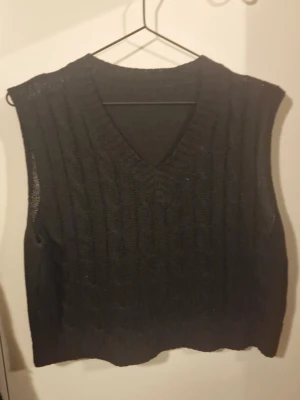 Svart stickad väst med v-ringning - Säljer en svart stickad väst med v-ringad hals och diskret kabelstickat mönster framtill. Perfekt att slänga över en skjorta eller t-shirt för en chill och stilren look. Västen har en klassisk passform och är ärmlös.