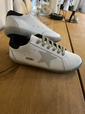 Golden goose - Säljer ett par vita Golden goose sneakers med grå mockadetaljer och en ikonisk stjärna på sidan. Skorna har grå snörning, svart hälparti och GGDB-logga i metall. Snygg kontrast mellan material och färger, perfekt till jeans eller kjol.