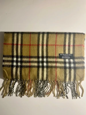 Rutig beige halsduk från Burberry - Klassisk halsduk från Burberry i 100% kashmir. Beige botten med ikoniskt rutigt mönster i svart, vitt och rött. Mjukt material och fransar i ändarna. Perfekt accessoar för att lyfta din outfit med en tidlös brittisk vibe.