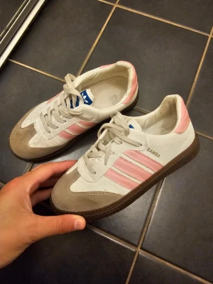 Adidas Samba sneakers med rosa detaljer - Säljer ett par Adidas Samba sneakers i vitt och beige skinn och mocka med klassiska rosa ränder på sidorna. Skorna har snörning och en brun gummisula med bra grepp. Perfekta för dig som vill ha en retro och sportig look med en touch av färg. Ej äkta