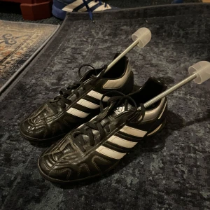 Svarta Adidas Puntero fotbollsskor - Säljer ett par klassiska Adidas Puntero fotbollsskor i svart med vita ränder. Skorna har snörning, rund tå och slitstark yttersula med bra grepp. Tillverkade i skinn och syntet för både komfort och hållbarhet. Perfekta för dig som vill dominera på planen.
