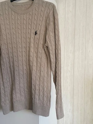 Beige kabelstickad tröja Ralph Lauren - Säljer en beige kabelstickad tröja från Ralph Lauren med klassisk rund halsringning och svart broderad logga på bröstet. Tröjan har lång ärm och ribbade muddar vid ärmslut och nederkant. Perfekt för lager på lager under hösten.