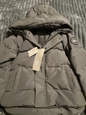 Mörkgrå pufferjacka från Canada Goose - Säljer en mörkgrå pufferjacka från Canada Goose äkta  med huva och diskret logga på ärmen. Jackan har dragkedja, knappar och två stora fickor framtill. Perfekt för kalla dagar och riktigt snygg passform.