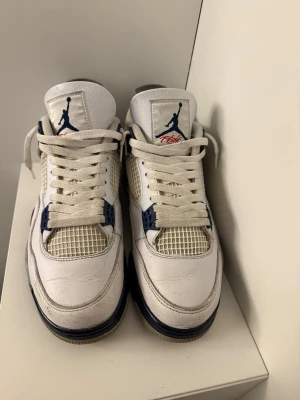 Nike Air Jordan 4 Retro Vit/Marinblå - Säljer ett par Nike Air Jordan 4 Retro i vitt och marinblått med meshpaneler, klassisk Jumpman-logga och detaljer i grått. Skorna har snörning, vadderad plös och sula med stänkmönster. Perfekta för dig som vill ha en ikonisk sneaker med streetwear-vibe.