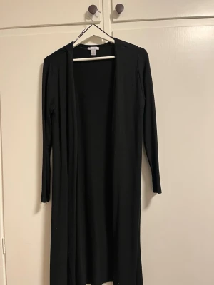 Svart lång kofta från Amisu - Lång svart kofta från Amisu med öppen front och långa ärmar. Enkel och stilren design som passar perfekt till lager på lager. Tillverkad i mjukt material som känns skönt mot huden.