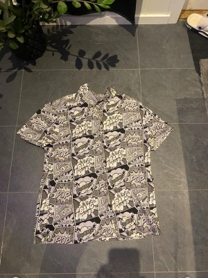 FSBN svartvit skjorta med serietidningsmönster - Cool skjorta från FSBN i svartvitt serietidningsmönster med ord som 'BANG', 'POP' och 'BOOM'. Skjortan har korta ärmar, klassisk krage och knäppning framtill. Regular fit och tillverkad i bomull, perfekt för dig som gillar statement-plagg.