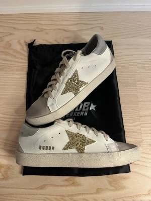 GGDB Superstar sneakers med glitterstjärna - Snygga GGDB Superstar sneakers i vitt skinn med beige snörning och silverfärgad häl. På sidan sitter en stor stjärna i guldglittrigt material som ger extra edge. 😊