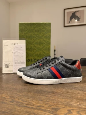 Gucci skor - Snygga Gucci sneakers i mörkgrått med GG-monogram, vita sulor och klassiska blå-röda ränder på sidan. Baksidan har metallicdetaljer i rött och blått ormskinnsmönster. Skorna har snörning och rund tå, tillverkade i canvas och skinn. Storlek 42