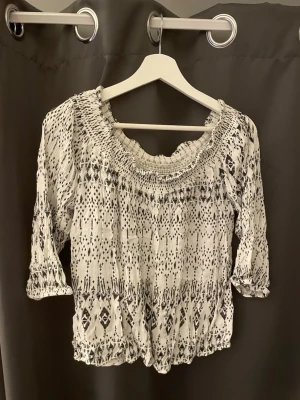 Vit och svart mönstrad blus  - Säljer en luftig vit blus från C i storlek S med svart grafiskt mönster. Blusen har bred resår vid halsringningen och trekvartsärm med resår vid ärmslut. Materialet är lätt och känns som bomull, perfekt för en avslappnad look.