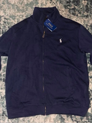 Marinblå kofta från Polo Ralph Lauren - Säljer en marinblå kofta från Polo Ralph Lauren med dragkedja framtill och klassisk vit logga på bröstet. Tröjan har hög krage, ribbade muddar och två fickor. Tillverkad i mjuk bomull som känns skön mot huden.