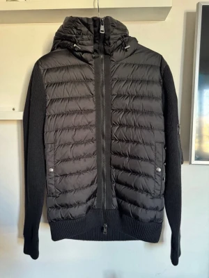 Moncler cardigan  - Säljer en svart hybridjacka från Moncler med quiltad front och huva i pufferstil. Ärmar och rygg är stickade för extra komfort och still. Säljer för den e för lite | 