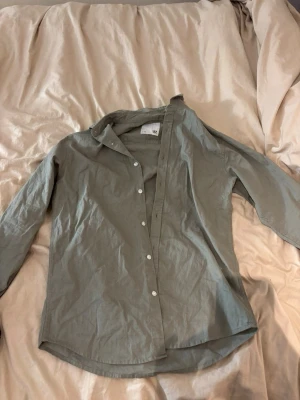 Grön skjorta - Säljer en stilren grå skjorta från 157 med klassisk krage och vita knappar framtill. Skjortan är långärmad och har en rak passform, perfekt att matcha med jeans eller chinos. Materialet känns lätt och skönt mot huden.