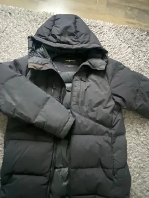 Svart pufferjacka Sail Antarctica - Säljer en svart pufferjacka från Sail Antarctica Expedition. Jackan har huva, dragkedja framtill och en patch på ärmen. Den är vadderad och riktigt varm, perfekt för kalla dagar. Klassisk passform och stilren look.