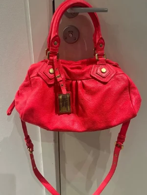 Röd axelväska från Marc by Marc Jacobs - Cool röd axelväska i skinn från Marc by Marc Jacobs med guldfärgade metalldetaljer och justerbar axelrem. Väskan har dubbla handtag, dragkedja upptill och ett snyggt innerfoder med logotypmönster. Den skiftar i färg lite beroende på vilket ljus man ser den i, men den är röd/neon röd rosa! Perfekt för dig som vill sticka ut eller söker efter en färgklick i din outfit!😇💞💞