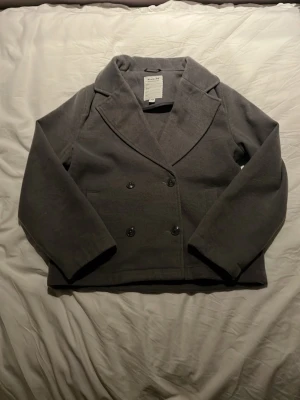 Grå vårjacka/höstjacka från Kappahl - Säljer en stilren grå vår/höstjacka från Kappahl. Jackan har dubbelknäppning med fyra knappar framtill, bred krage och raka ärmar. Perfekt för dig som gillar klassisk och enkel design.