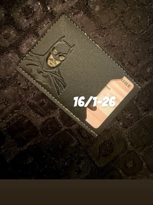 Svart Batman plånbok i skinn - Säljer en svart plånbok med Batman-motiv i relief. Plånboken har plats för kort och är tillverkad i slätt skinn med vita sömmar runt kanterna. Perfekt för dig som gillar superhjältar och vill ha något unikt i fickan.