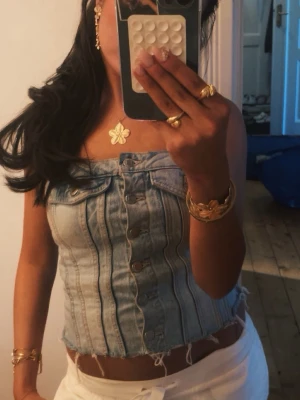 Ljusblå denim korsettopp - Trendig ljusblå korsettopp i denim med markerade sömmar och knappar framtill. Toppen är croppad och har råa kanter nedtill för en edgy look. Perfekt att styla med smycken och höga byxor.