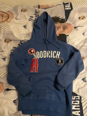 Blå hoodie från Hoodrich med patchar - Blå hoodie från Hoodrich med vita och röda detaljer, broderade patchar och stor H på bröstet. Tröjan har huva med snörning, känguruficka och ribbade muddar. Perfekt för dig som gillar streetwear och vill sticka ut med coola detaljer.