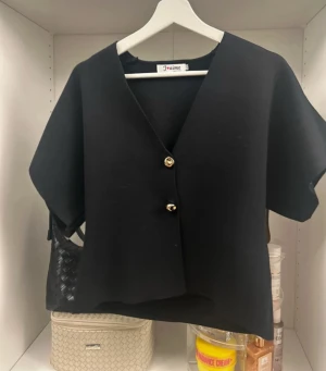 Svart bolerotopp  - Säljer en svart bolerotopp  med v-ringning och två snygga guldfärgade knappar framtill. Toppen har korta ärmar och är tillverkad i ett mjukt, stickat material som ger en chill och modern vibe.