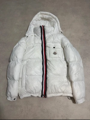 Vit Moncler pufferjacka med huva - Säljer en fet vit pufferjacka från Moncler med huva och klassisk logga på bröstfickan. Jackan har dragkedja framtill med röd, vit och blå rand, stora fickor och justerbar nederkant. Perfekt för kalla dagar när du vill hålla stilen.