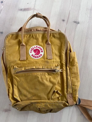 Gul Kånken ryggsäck från Fjällräven - Klassisk Kånken ryggsäck från Fjällräven i en snygg gul färg. Väskan har det ikoniska runda logotypmärket framtill, dragkedja, framficka och justerbara axelremmar. Tillverkad i slitstarkt tyg och har praktiska handtag upptill.