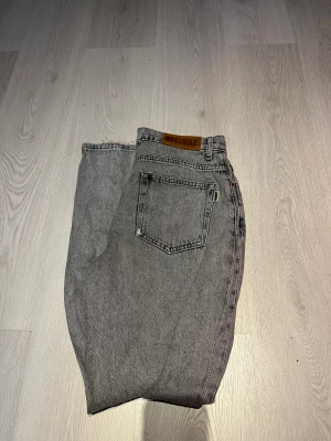 Grå jeans från Woodbird med rå kant - Säljer ett par grå jeans från Woodbird med rak passform och rå, fransig kant vid bensluten. Klassisk femficksdesign och snyggt läderpatch med logga bak. Jeansen har en cool, lite tvättad look och är tillverkade i slitstarkt bomullsmaterial.