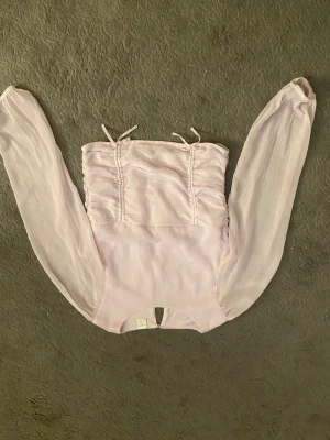 Ljuslila blus med långa ärmar från Lindex - Supersöt ljuslila blus från Lindex med långa, transparenta ärmar och smockad rygg. Toppen har knytband upptill och rynkade detaljer framtill. Perfekt för dig som gillar romantisk och feminin stil.