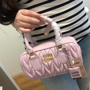 Ljusrosa quiltad handväska från Miu Miu - Supersöt ljusrosa handväska från Miu Miu med quiltad design och guldfärgade detaljer. Väskan har ett stilrent format, dubbla handtag och en snygg dragkedja upptill. Perfekt för dig som gillar lyxiga accessoarer med en feminin touch.