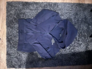 Mörkblå hoodie Peak Performance - Säljer en mörkblå hoodie från Peak Performance med dragkedja framtill och broderad logga på bröstet. Tröjan har huva. Det står ingen direkt storlek bara storlek 160 men den är lika stor som en xs 