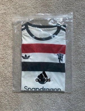 Manchester United x Adidas fotbollströja - Snygg Manchester United fotbollströja från Adidas med breda ränder i vitt, svart och rött. Tröjan har korta ärmar, svart krage och detaljerade loggor på bröstet och ärmarna. Materialet är lätt och andas, perfekt för match eller träning. Snapdragon sponsortryck på framsidan.
