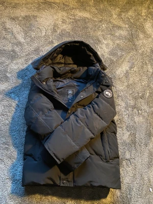 Svart pufferjacka från Canada Goose - Svart pufferjacka från Canada Goose med huva och klassisk patch på ärmen. Jackan har en stilren look, raka linjer och är fodrad för extra värme. Perfekt för kalla dagar och har dragkedja samt tryckknappar framtill. Snygg och funktionell för vintern. Sista bilden ser man den på!