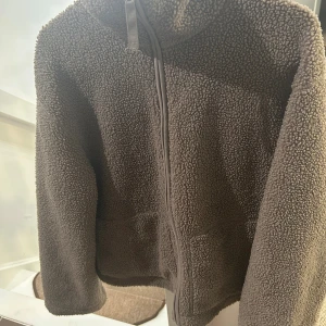 Brun teddyjacka med dragkedja - Mysig brun teddyjacka med huva och hel dragkedja framtill. Jackan har en fluffig och mjuk yta som ger en avslappnad vibe. Perfekt för kyliga dagar när du vill vara både varm och stilren.