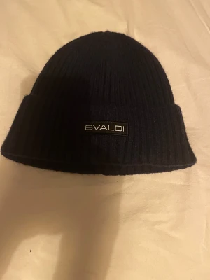 Marinblå ribbad mössa från Bvaldi - Marinblå ribbstickad mössa från Bvaldi med uppvikt kant och diskret logotyp framtill. Klassisk form som sitter snyggt på huvudet och passar perfekt när det är kallt ute. Tillverkad i mjukt material som håller värmen. 