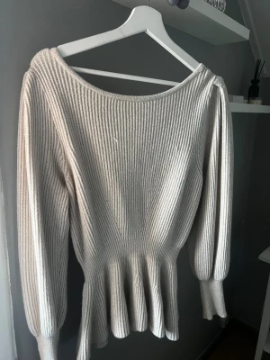 Beige ribbad stickad tröja med volang - Säljer en beige stickad tröja med ribbad struktur och snygg volangkant nedtill. Tröjan har långärmade puffärmar och bred rund halsringning. Perfekt för dig som gillar en feminin och trendig look.
