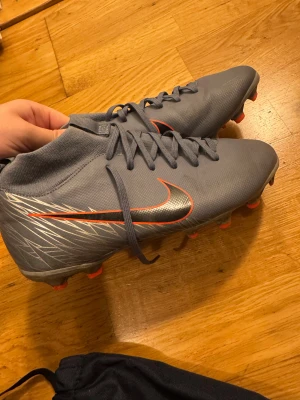 Grå Nike fotbollsskor med röd detalj - Säljer ett par grå Nike fotbollsskor med snygga röda och silverfärgade detaljer på sidan. Skorna har snörning och platt sula med dobbar för bra grepp på planen. Materialet är syntet och designen är modern med en sportig look.