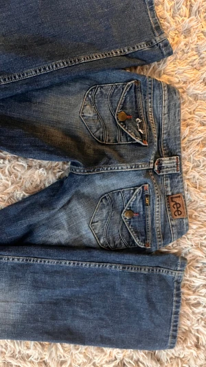 Blå bootcut jeans från Lee - Snygga blå lågmidjade  jeans från Lee med bootcut passform och slitna detaljer på benen. Jeansen har coola fickor med lock och knappar, samt Lee-logga på bakfickan. Materialet är klassisk jeans och färgen är mörkblå med tvättade partier. Perfekt för dig som gillar en avslappnad och trendig look. Lite slitna längst ner inga bilder på