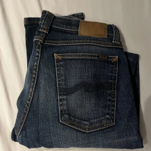 Mörkblå jeans från Nudie Jeans - Snygga mörkblå jeans från Nudie Jeans med klassisk femficksdesign och kontrastsömmar. Jeansen har en rak passform och är tillverkade i slitstarkt denim med diskreta slitningar för en cool look. Perfekta för dig som gillar stilrena och tidlösa jeans.W27 L32