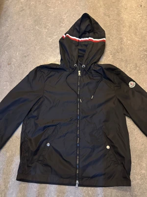 Svart vindjacka från Moncler med huva - Svart vindjacka från Moncler med huva och dragsko, dragkedja framtill och två fickor med tryckknappar. Snygg detalj med röd och vit rand på huvan samt Moncler-logga på ärmen. Perfekt för dig som vill ha en clean och sportig look.