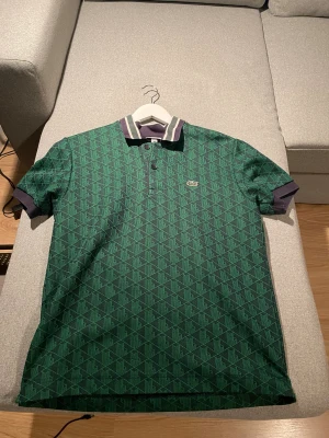 Grön mönstrad piké från Lacoste - Snygg grön pikétröja från Lacoste med geometriskt mönster i mörkgrönt och svart. Klassisk krage med vita och gröna ränder, samt knappar framtill med Lacoste-logga. Tröjan har korta ärmar och är i klassisk passform. Perfekt för dig som gillar sportig och stilren look.