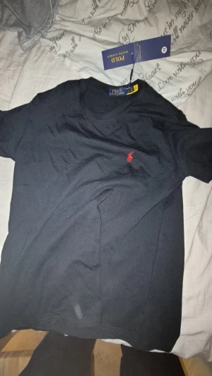 Svart t-shirt från Polo Ralph Lauren - Svart t-shirt från Polo Ralph Lauren i mjuk bomull. Klassisk rund hals och korta ärmar. På bröstet finns den ikoniska röda Polo-loggan broderad. Perfekt basic med stilren look och snygg passform.