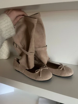 Beige ballerinaskor med unik design - Säljer ett par snygga beige ballerinaskor i mocka med rund tå och söt rosettdetalj framtill. Skorna har en platt sula och är perfekta för dig som gillar en enkel men chic stil. Passar till både jeans och klänning.