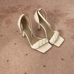 Vita klackskor med flätad rem✨💖✨ - Säljer ett par vita sandaler med hög klack från Bianco. Skorna har fyrkantig tå, flätad rem över tårna och justerbart ankelband med spänne. Tillverkade i syntetmaterial med stilren och modern look, perfekta för att lyfta din outfit 🫶🏻🫶🏻🫶🏻